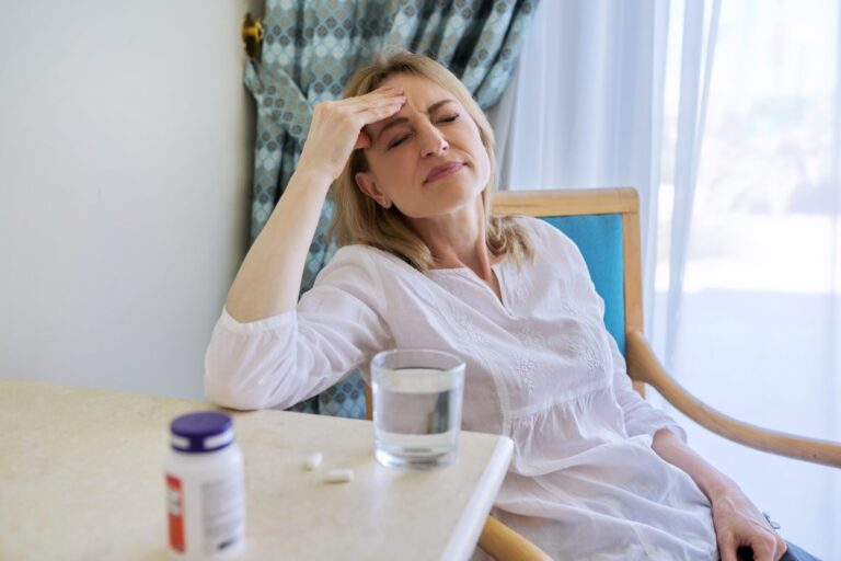 Woman at a table holding her forehead beside a pill bottle and glass of water