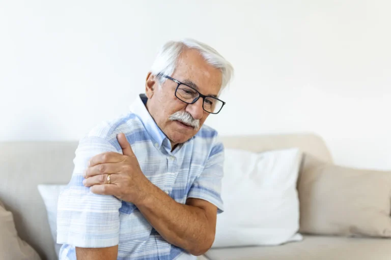 Older man seated on a sofa holding his shoulder in pain