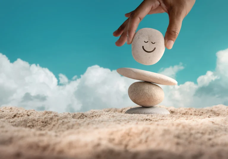 Hand placing a smiling pebble on top of balanced stacked stones against a bright sky