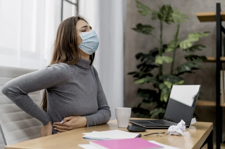Woman in a mask at a desk holding her lower back