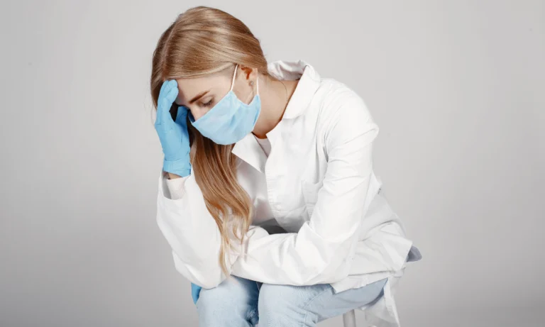 Masked clinician in a white coat holding her head in fatigue