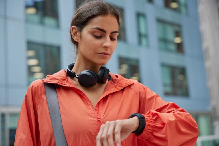 Woman in a jacket and headphones checking a smartwatch outdoors