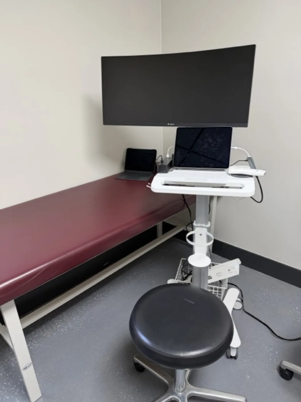Point-of-care ultrasound system with a handheld transducer resting in its cradle on a mobile workstation, cabled to a tablet, with a wall-mounted display beside the examination table, at Padda Institute, 4477 Woodson Road, St. Louis, MO 63134