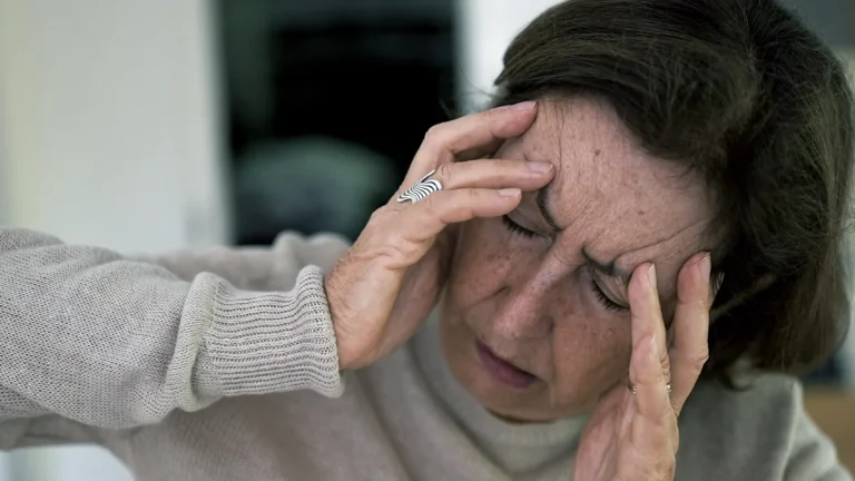 Older woman seated indoors holding her forehead