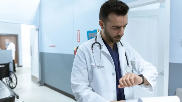 A doctor in a white lab coat checks his wristwatch in a hospital hallway, conveying professionalism.