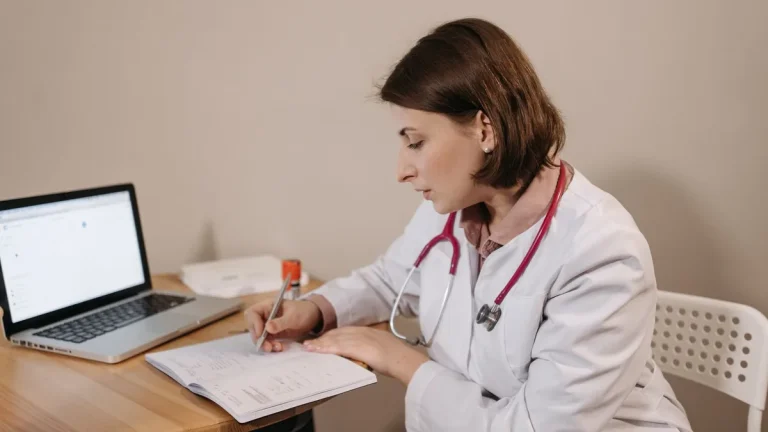 A female doctor in a lab coat writing a prescription in a clinic office setting with a laptop on the table.