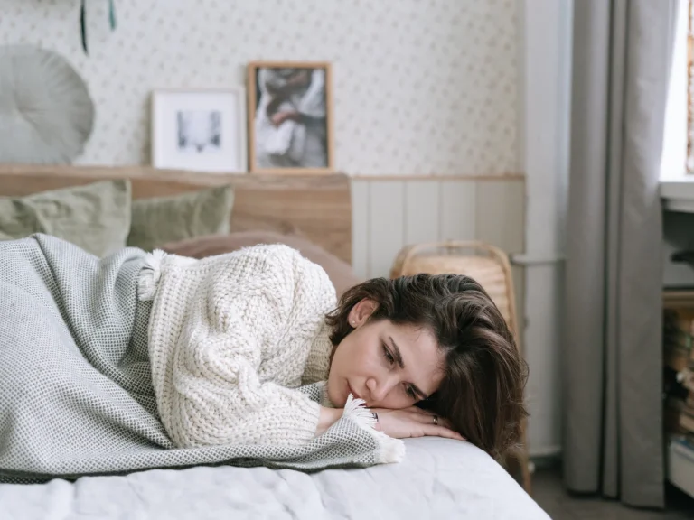 A person lying awake in bed under a blanket during the day, eyes open