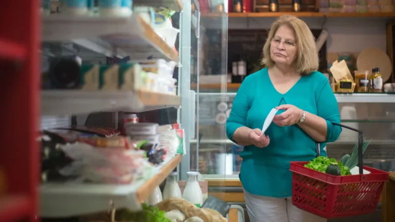 An older woman with a shopping basket and list in a grocery store aisle