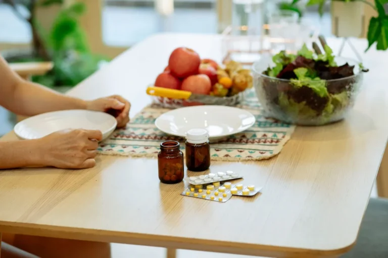 Pill bottles and blister packs on a dining table beside a bowl of leafy salad and a bowl of fruit