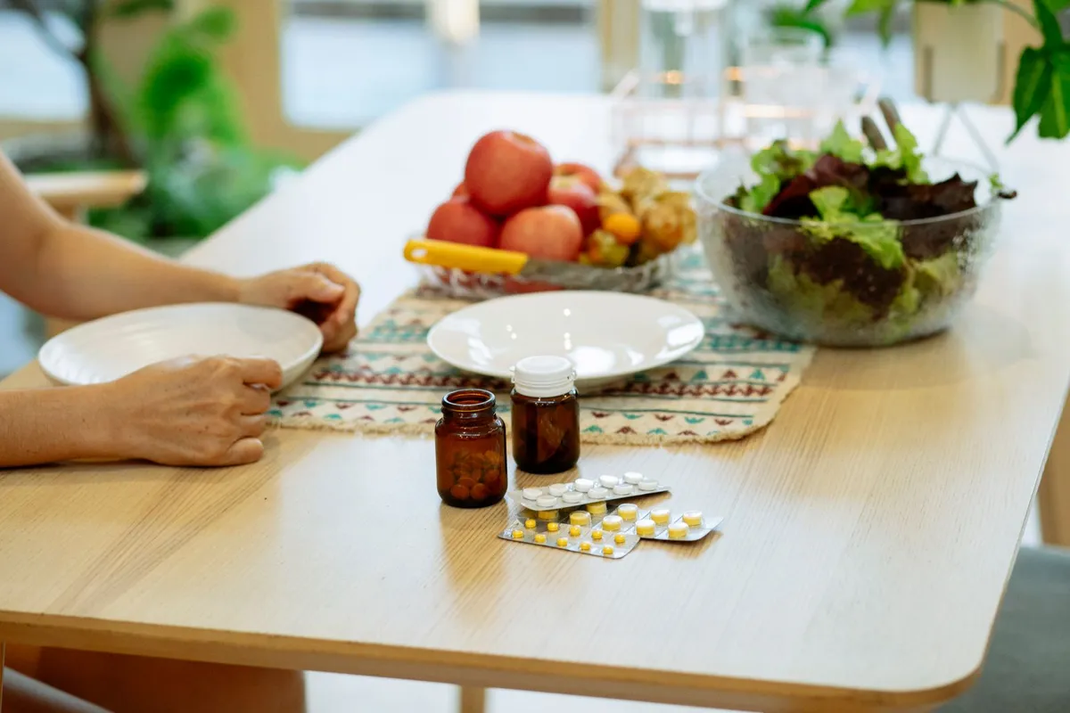Pill bottles and blister packs on a dining table beside a bowl of leafy salad and a bowl of fruit